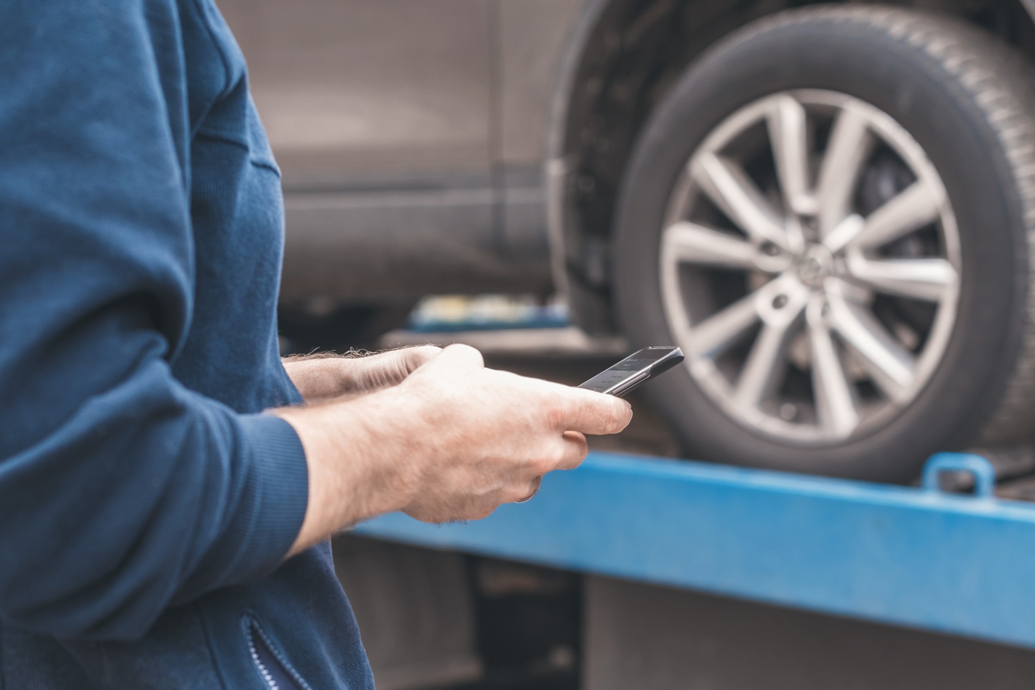 A person in a blue hoodie is holding a smartphone next to a car being towed on a flatbed truck. The image focuses on the person's hands and the car's wheel in the background.