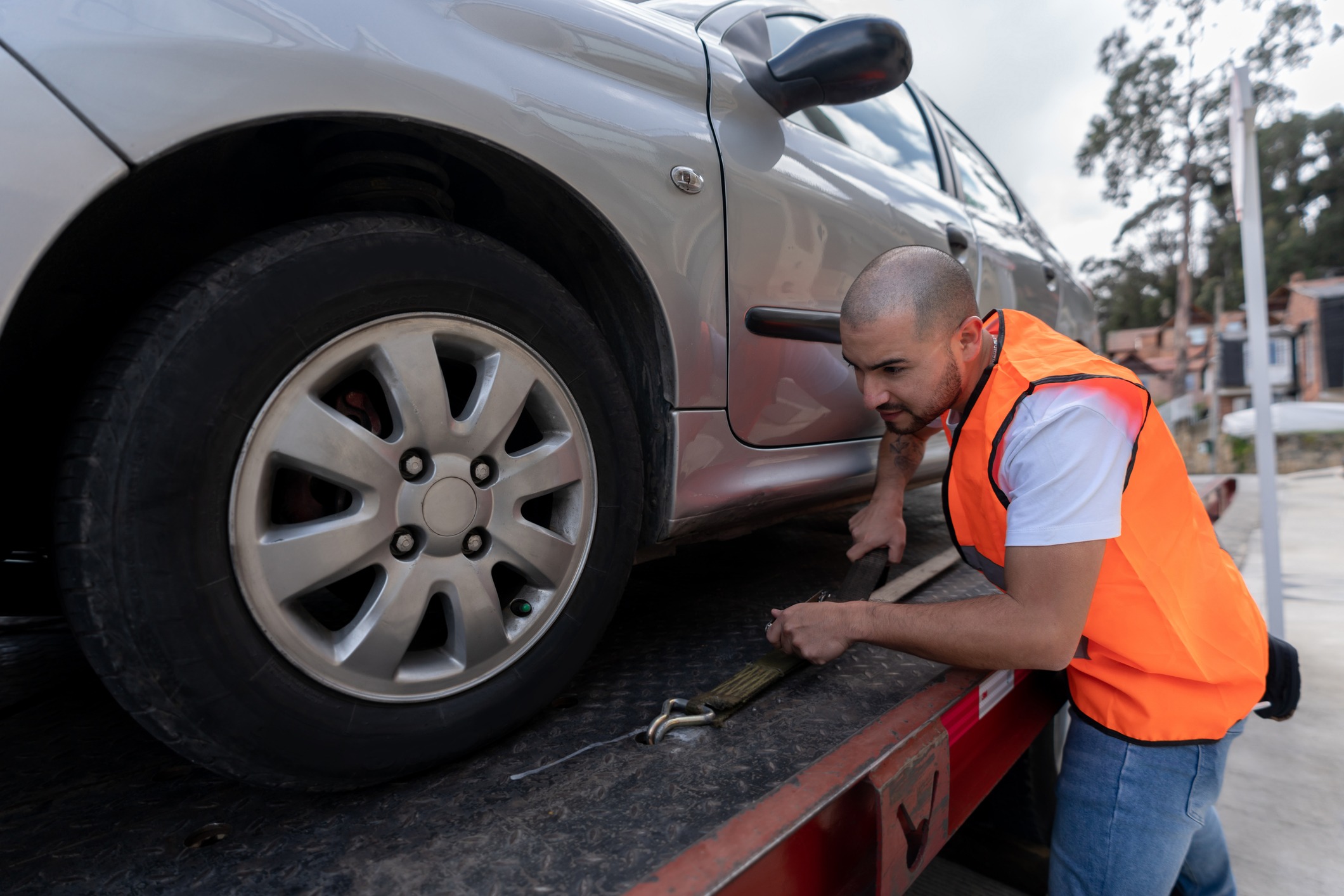 A man in an orange safety vest is securing a gray car onto a tow truck using a strap. The car is on a flatbed, and the scene is outdoors on a cloudy day. Trees and buildings are visible in the background.